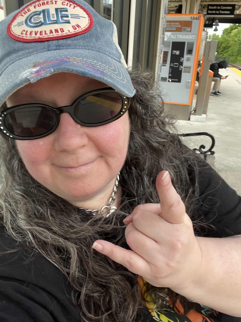 A selfie of Christine who is wearing studded sunglasses and a jean baseball cap repping Cleavland Ohio. She's holding up her left hand in the Rock On gesture. 