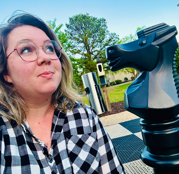 Kel is wearing a black and white checkered shirt and glasses while looking pensive. Their eyes are looking up and to the right. Behind them and to the right is a Black Knight chess piece on a human-sized chess board.