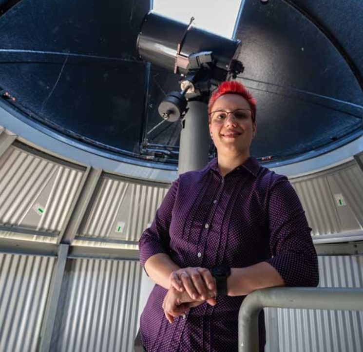 Skiiz has bright red hair and a purple button down shirt. She's standing inside an observatory with a large telescope behind her.